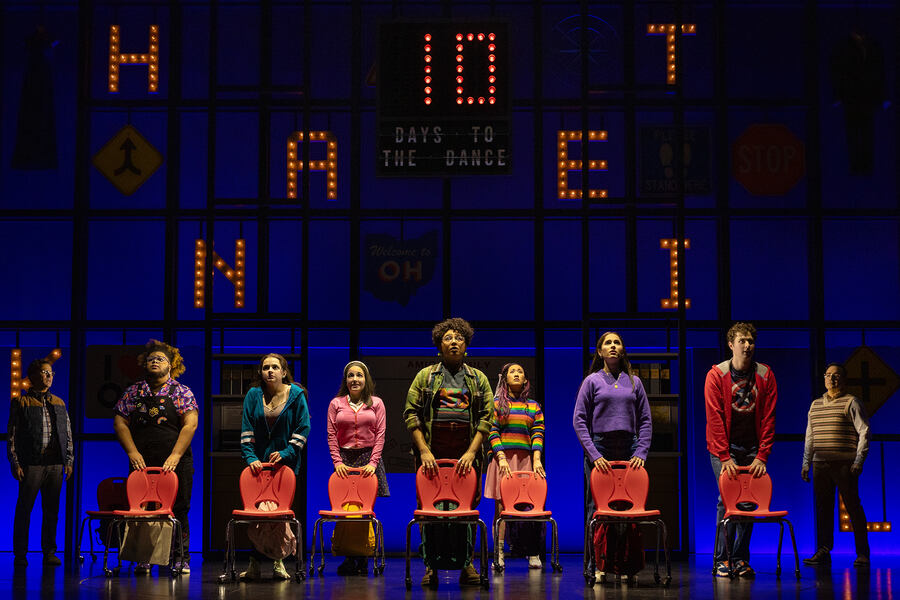 The seven main characters of 'How to Dance in Ohio' stand in a line across the stage behind red classroom chairs, wearing brightly-colored, contemporary teen clothing. Two teachers stand on either side. There is a blue, backlit background with a grid pattern, adorned with an assortment of neon-bulbed letters. At center, a sign reads “10 days to the dance.”