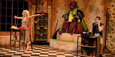 A production image in which a blonde white leading lady sings, her arms outstretched, on the set of a flower shop; behind her to left is a white man dressed as a giant plant, and far stage left, a bespectacled young white man, looking nervously, holds up a telephone receiver.