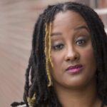 A headshot of Kayla Hamilton, who is a dark brown-skinned Black woman with black and golden highlighted dreads. She is posing in front of a blurred brick wall.