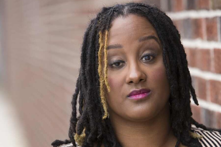 A headshot of Kayla Hamilton, who is a dark brown-skinned Black woman with black and golden highlighted dreads. She is posing in front of a blurred brick wall.