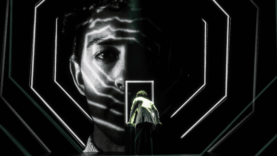 At center stands a steel table and glowing white frame, meant to represent a futuristic pinball machine. Adult Tommy wears a white blazer and is playing pinball with his back to the audience. A massive black and white projection of his face, framed in white neon hexagonal lights, stares out from the dark background.