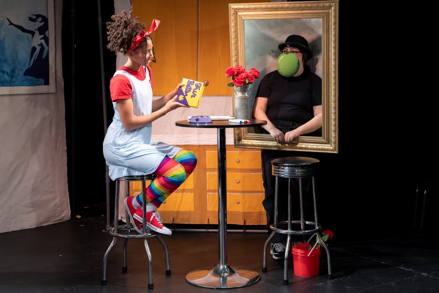 The image depicts a whimsical theater scene with two performers. On the left, a lively character in colorful attire, including rainbow leggings and a red bandana, sits on a stool next to a high-top table and holding a small box of cat food. On the right, a performer dressed in black and wearing a bowler hat stands behind a gold picture frame. Their face is obscured by an image of a green apple, mimicking René Magritte's The Son of Man painting. The high-top table between them is adorned with flowers and art supplies.