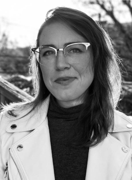 Black-and-white image of a white femme with medium-length hair looking into the camera. She wears glasses, a turtleneck, and a white jacket. In the background, there are leafless trees.