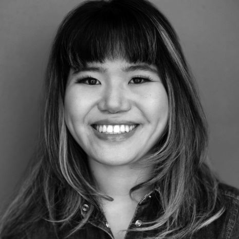 A black-and-white image of a Chinese, Jewish, transracial femme looking into the camera with a bright smile. Her long hairstyle features bangs and highlights, and she wears a denim jacket. The background is plain.