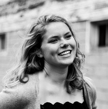 A black-and-white image of a white femme wearing a cardigan and top with a scalloped neckline. She smiles to the side of the camera, and her hair flows in the wind. There is a building out of focus in the background.