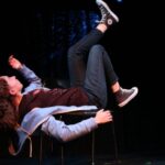 Sydney, a white young person with shoulder length light brown curly hair wearing a black NASA t-shirt, blue jeans, black converse, and a light blue zip up hoodie, is laying on their back on a chair with their arms and legs in the air as if they are an astronaut. The lighting is mostly dark with some blue speckles around them.