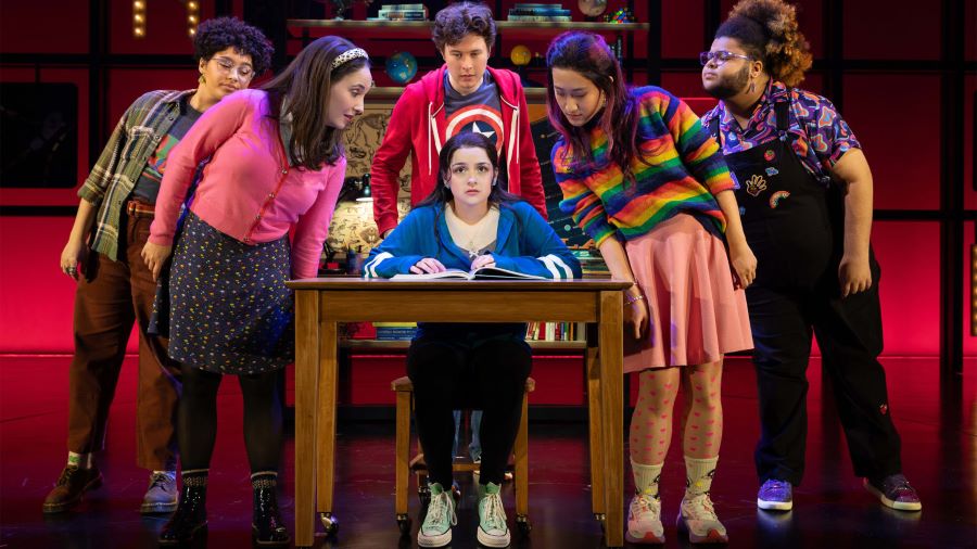 A color performance image of Broadway's HOW TO DANCE IN OHIO. A young femme sits at a desk with paper, looking out at the audience and wearing a bright blue jacket. Five of the characters' friends peer over at the paper on her desk. They wear colorful clothing, including a Captain America shirt, a rainbow top, and other items with small dotted or squiggly designs.