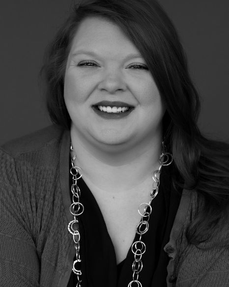 A black-and-white image of a white femme with long hair. Wearing a bold lipstick, long necklace, cardigan, and v-neck, she beams into the camera. The background is plain.