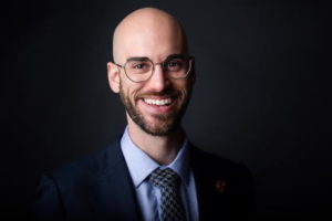 A man, bald on top but with a beard and moustache, in a studio portrait. He is wearing glasses and a suit and tie.